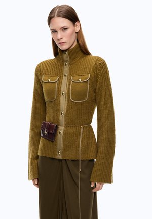 Mujer con chaqueta de punto verde oliva con bolsillos abotonados, cuello alto, un cinturón de cadena y una pequeña bolsa marrón oscuro pegada a la cintura.