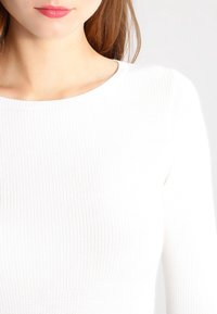 Weißes, geripptes Langarmshirt mit rundem Ausschnitt. Das Material wirkt weich mit leichter Textur, körperbetonter Schnitt und nahtlose Schultern.