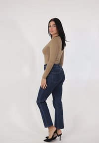 Top de manga larga ajustado de color beige con jeans de tiro alto azul oscuro. Tacones de aguja negros con tiras transparentes. Fondo neutro.