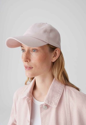 Vrouw met lang blond haar die een lichtroze honkbalpet en bijpassende jas draagt, kijkt iets naar links tegen een effen achtergrond.