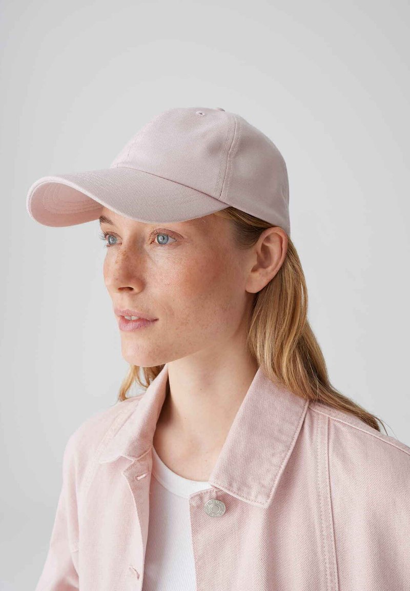 Frau mit langen blonden Haaren, die eine hellrosa Baseballkappe und eine passende Jacke trägt, schaut leicht nach links vor einem einfarbigen Hintergrund.