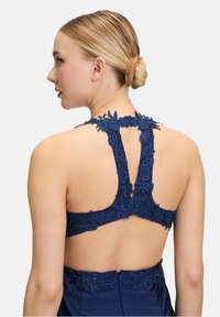 Granatowa sukienka z kwiatowym koronkowym wykończeniem na plecach, prezentująca bezrękawowy krój oraz odkryte plecy z dekoracyjnymi paskami i gładką teksturą materiału.