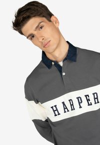 Polo a maniche lunghe grigio con colletto blu scuro e striscia orizzontale bianca che presenta il nome "HARPER" ricamato in navy. Tessuto morbido.