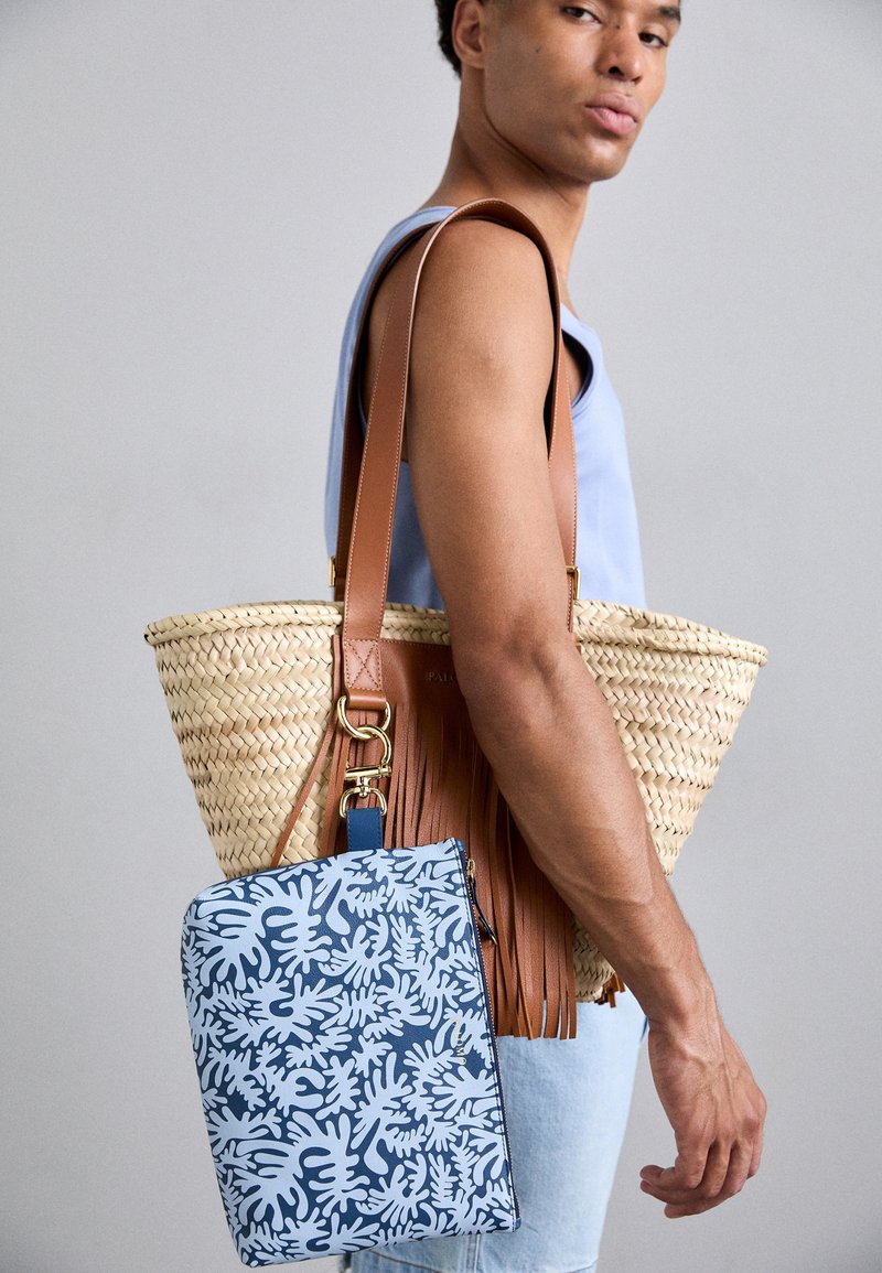 Bolso de paja tejido con correa de cuero marrón y flecos; pouch azul con diseño abstracto colgando del bolso.