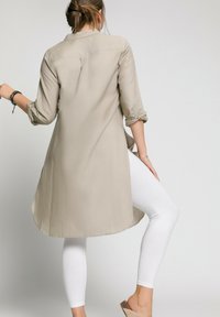Chemise longue beige clair avec des manches retroussées, détails de boutons et un ourlet incurvé, associée à un legging blanc et des sandales beige.