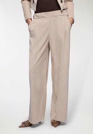Pantaloni a gamba larga in corduroy beige con cintura elastica, tasche laterali e texture a righe verticali sottili. Abbinati a scarpe eleganti marroni a punta.