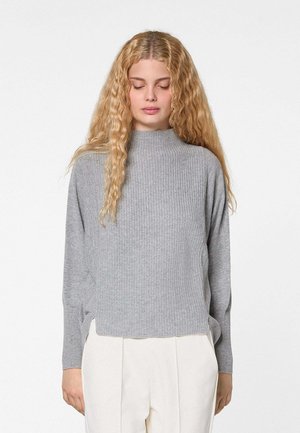 Vrouw met lang, golvend blond haar, gekleed in een lichtgrijze geribbelde trui en witte hoge taillebroek, staand met gesloten ogen tegen een effen achtergrond.