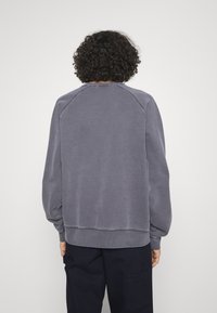 Persona con cabello rizado, vistiendo una sudadera gris de manga larga y holgada, y pantalones oscuros, de pie de espaldas frente a un fondo plano.
