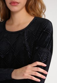 Czarna bluzka z długim rękawem o teksturowanym wzorze z motywem chevron. Tkanina ma subtelną połyskliwość i okrągły dekolt.