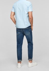 Camicia azzurro chiaro a maniche corte con un motivo a pois, abbinata a jeans blu scuro con risvolti e sneakers bianche. Visibile da dietro.