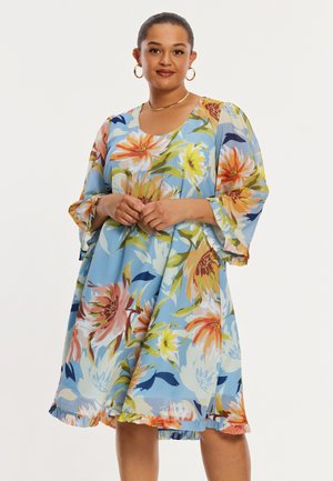 Femme portant une robe bleue mi-longue à grands motifs floraux colorés, des boucles d'oreilles anneaux dorées et un collier en or, debout les mains jointes.