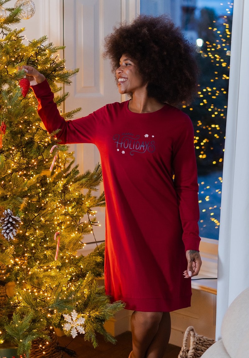 Rode nachtkleding met lange mouwen met de tekst "Happy Holidays" in grijs, gedragen door een persoon naast een versierde kerstboom met lichtjes.