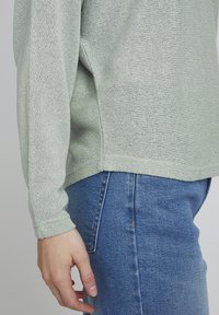 Hellgrüner, strickpullover mit langen Ärmeln und seitlichen Schlitzen, kombiniert mit blauen Jeans. Strukturiertes Material mit lockerer Passform.