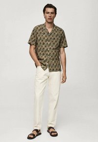 Kortærmet skjorte med et geometrisk mønster i grøn, brun og creme, parret med cremefarvede bukser og sorte sandaler.