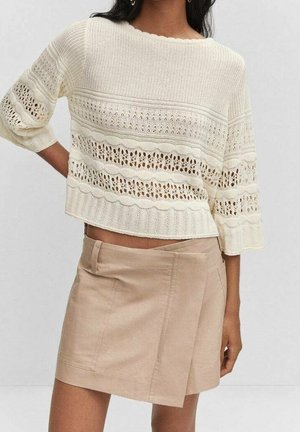 Femme portant un pull court en maille crème avec des motifs en dentelle et une mini-jupe asymétrique beige, debout devant un fond uni.