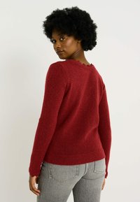 Suéter rojo con un acabado texturizado, mangas largas y detalle de escote con volantes, combinado con unos jeans grises.