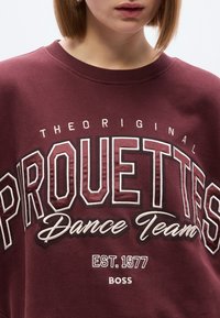 Μπορντό φούτερ με την επιγραφή «PROUETTES Dance Team» σε λευκό χρώμα, με λεπτομέρειες όπως «EST. 1977» και «BOSS» σε μικρότερο κείμενο.