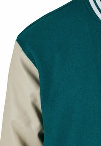 Hanorac din țesătură teal, cu un manșon din piele bej neted. Prezintă un guler cu nervuri, cu dungi gri și albe. Textură moale în general.