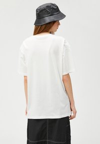 Camiseta blanca de gran tamaño en algodón, con mangas cortas y cuello redondo, combinada con un sombrero tipo cubo negro. Diseño sencillo, sin estampados.