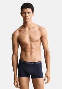 Junger Mann steht, trägt marineblaue GANT Boxershorts, oberkörperfrei, schaut geradeaus, schlichter heller Hintergrund.