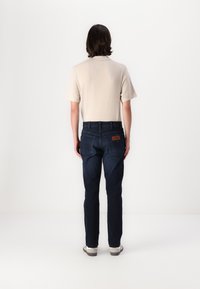 Beige, rövid ujjú ing, sötétkék farmernadrággal párosítva. A farmernadrágnak van egy barna bőrpaskája és két hátsó zsebe. A modell háttal áll.