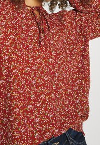 Blusa rossa con piccolo motivo floreale rosa, con scollo annodato, indossata con jeans blu scuro, capelli ricci parzialmente visibili.