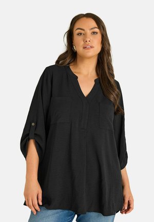 Vrouw met lang bruin haar die een losse zwarte blouse draagt met opgerolde mouwen en lichtblauwe spijkerbroek, staand tegen een witte achtergrond.