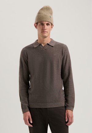 Brun langærmet polo skjorte med v-hals og ribbet tekstur. Parret med mørkebrune bukser. Modellen bærer en sandfarvet strikket beanie.