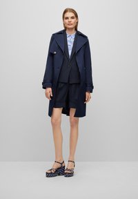 Tumesinine trench coat pinstripi bleiseri ja lühikeste pükste komplekti peal, millele on sobitatud tumesinised täpilised platvormi sandaalid. All on helesinine kraega särk.