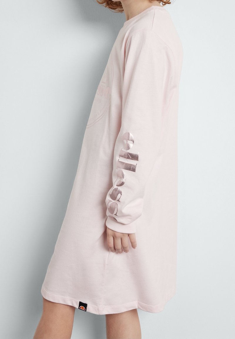 Enfant portant une robe rose clair à manches longues avec le texte métallique "ellesse" sur la manche et une petite étiquette noire avec logo près de l'ourlet, vue de profil.
