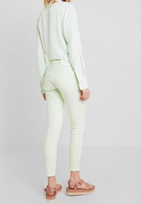 Jeans ajustados de color verde claro con bolsillos traseros, combinados con una blusa de color menta y sandalias de tira rosa sobre un fondo claro. Textura suave.