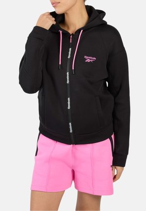 Vrouw draagt een zwart Reebok-hoodie met ritssluiting, roze trekkoorden en logo, gecombineerd met felroze sportshorts.