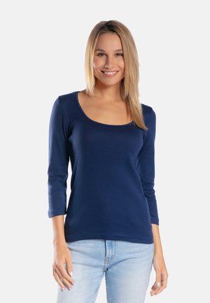 Uśmiechnięta kobieta o prostych blond włosach, ubrana w granatową bluzkę z długim rękawem i dekoltem w łódkę oraz jasnoniebieskie dżinsy, stojąca na białym tle.