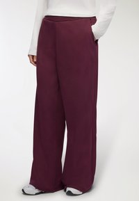 Spodnie o szerokich nogawkach w głębokim burgundowym kolorze, wykonane z gładkiego materiału, z elastycznym pasem i kieszeniami po bokach. Noszone z białymi trampkami.