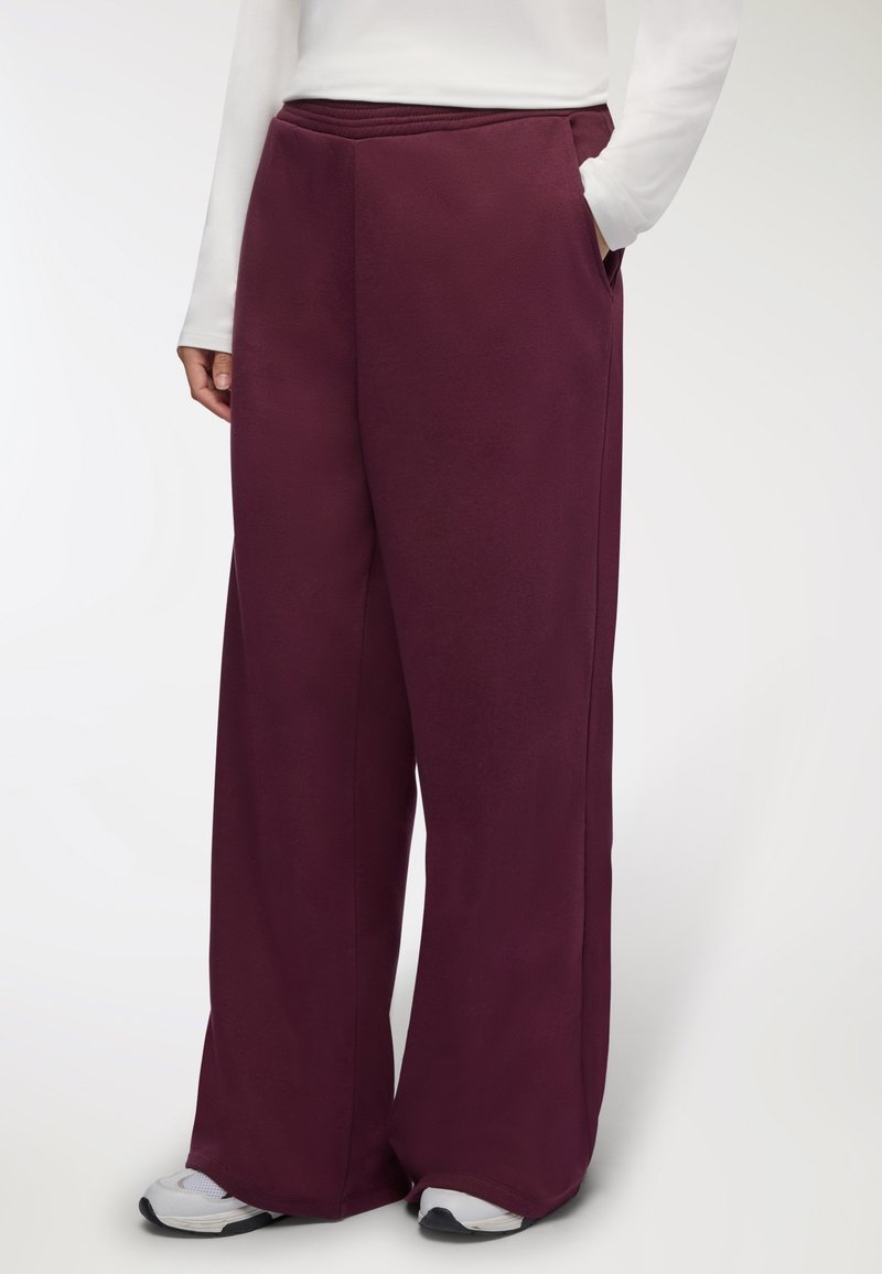 Spodnie o szerokich nogawkach w głębokim burgundowym kolorze, wykonane z gładkiego materiału, z elastycznym pasem i kieszeniami po bokach. Noszone z białymi trampkami.