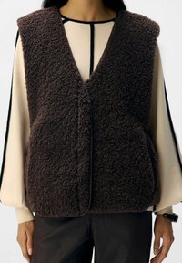 Gilet marrone morbido con una superficie testurizzata, progettato senza maniche, con scollatura rotonda e chiusura frontale.