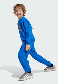 Sweatshirt bleu et joggeurs assortis en polaire, arborant le logo Adidas. Des baskets grises avec des rayures blanches complètent la tenue.