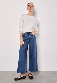 Witte gebreide trui met kabelpatroon, gecombineerd met een broek van blauwe denim met wijde pijpen. Model draagt zwarte mesh flats. Soepele, lichte textuur.