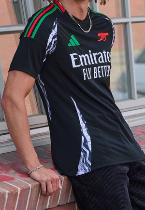 Person iført sort Arsenal fodboldtrøje med røde, grønne og hvide detaljer og "Emirates Fly Better" sponsorlogo, stående ved en murstenskant.
