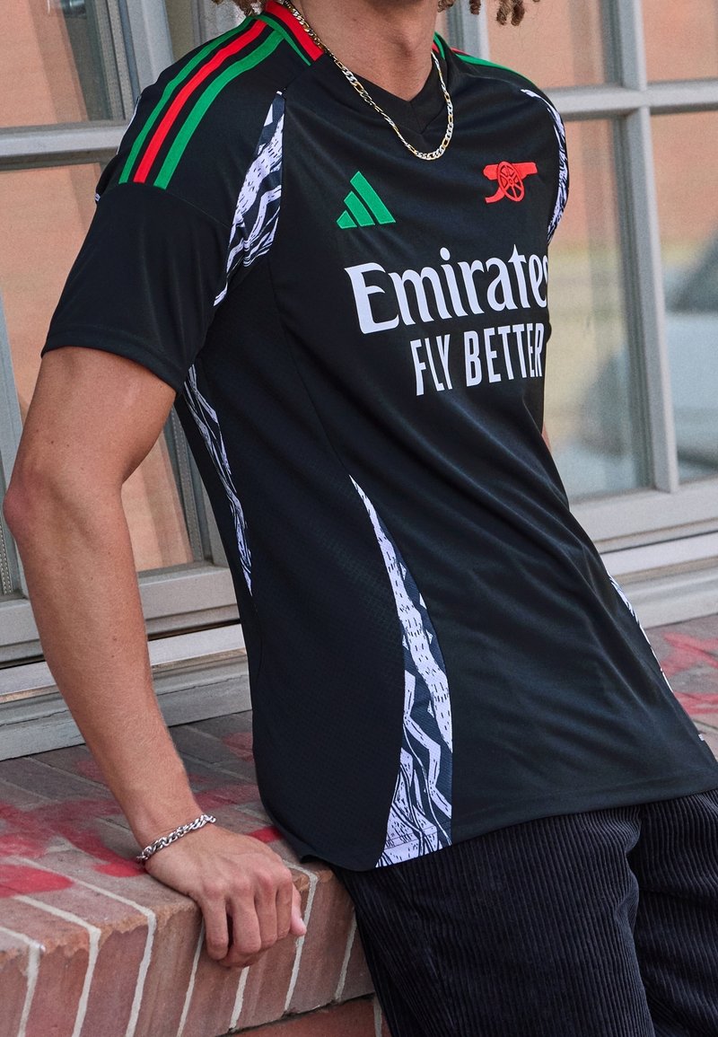 Personne portant un maillot de football noir d'Arsenal avec des détails rouges, verts et blancs et le logo du sponsor "Emirates Fly Better", debout près d'un rebord en briques.