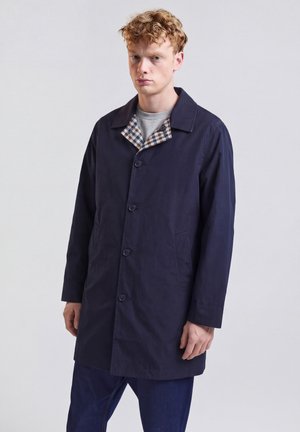 Junger Mann mit lockigem Haar, der einen dunkelblauen, geknöpften Mantel mit kariertem Innenkragen über einem grauen Hemd trägt und vor einem schlichten hellen Hintergrund steht.