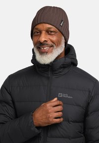 Zwarte geïsoleerde jas met een ritssluiting aan de voorkant en een logo op de borst, gecombineerd met een bruine geribbelde gebreide beanie. Glad textuur, warm ontwerp.
