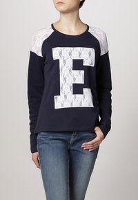 Felpa navy con dettagli in pizzo bianco sulle spalle e logo 'E' testurizzato bianco sulla parte anteriore. Realizzata in tessuto morbido e elastico.