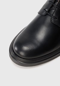 Nærbillede af en poleret sort læder pottesko tå med sorte snørebånd på en hvid baggrund.