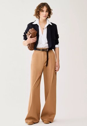 Giovane donna con pantaloni beige larghi, camicia bianca, giacca corta nera, che tiene una borsa marrone in pelle su uno sfondo semplice.