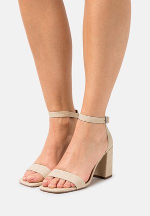 Sandales à talons bloc beiges avec une finition en simili cuir texturé. Elles possèdent une large bride sur les orteils et une bride de cheville réglable.