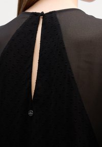 Vue arrière d'une personne portant un haut en tissu texturé noir avec une petite étiquette métallique circulaire et une ouverture en forme de trou de serrure verticale au niveau du haut du dos.