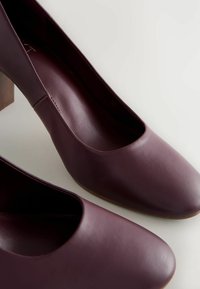 Burgundy nahkaiset korkokengät, joissa on sileä pinta, pyöreä kärki ja keskipituinen lohkokorko. Minimalistinen muotoilu ilman koristeluja.