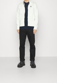 Man i vit zip-up jacka över en marinblå skjorta, svarta byxor och svarta sneakers som står mot en enkel vit bakgrund.