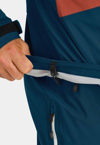Teal outdoor jacket with orange accents, showing a hand gripping the adjustable hem featuring a plastic clip and drawcord. Textured fabric.